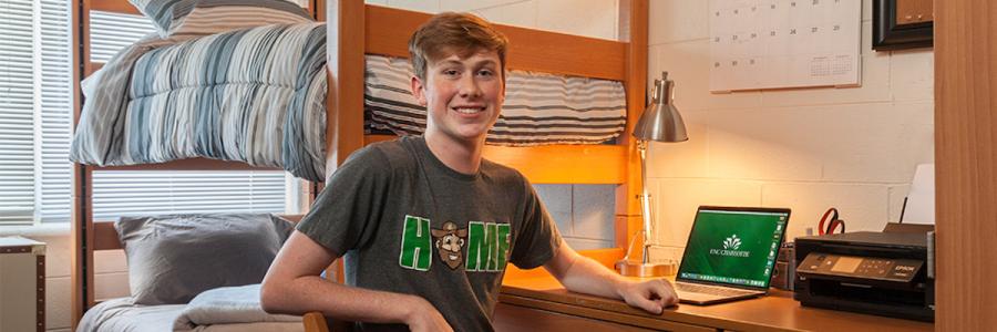 A new student shown smiling in front of dorm room desk