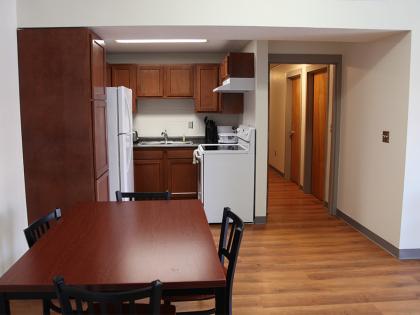 A room showing of a dining space consisting of a table with 4 chairs and a cooking area with a fridge, sink and stove
